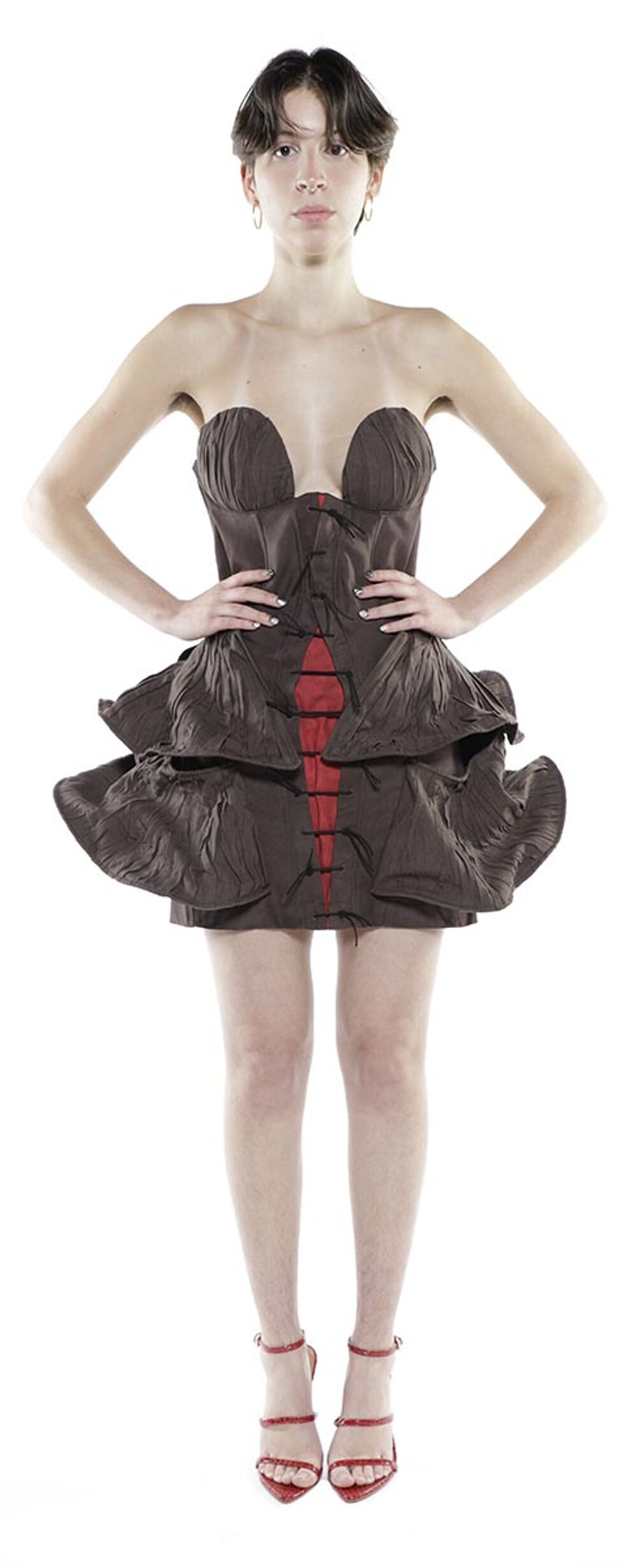 A woman with short brown hair stands facing forward wearing a brown corseted dress with red detailing and red high-heeled sandals.