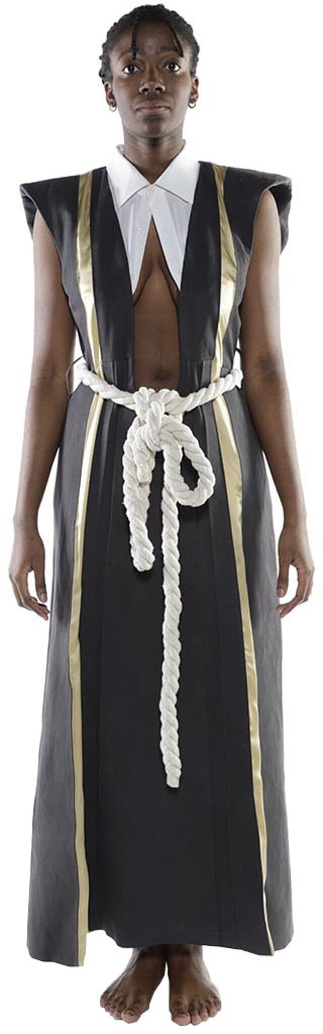 A dark-skinned person with short hair is standing, wearing a long, dark sleeveless robe with gold accents, a white collared shirt underneath, and a thick white rope tied around their waist.