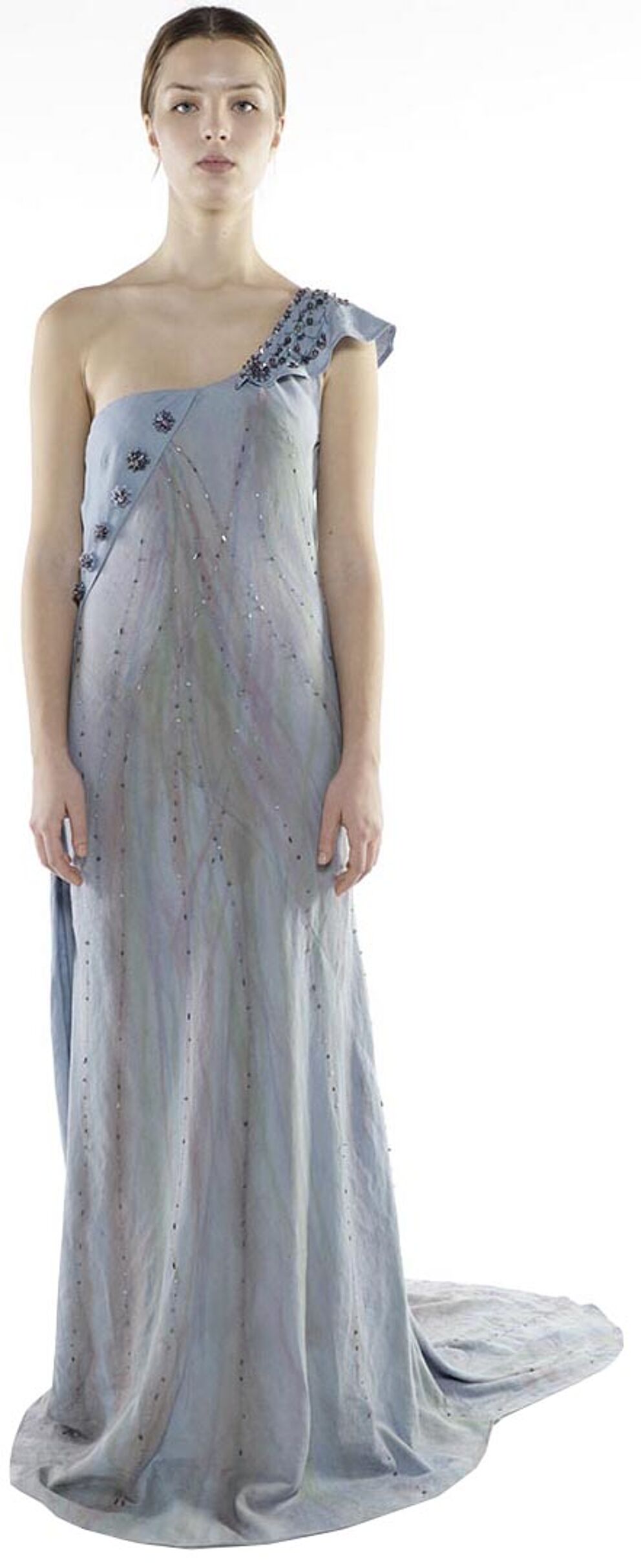 A slender woman with light skin and dark hair stands against a white background wearing a light-blue, one-shoulder, floor-length gown adorned with dark embellishments.