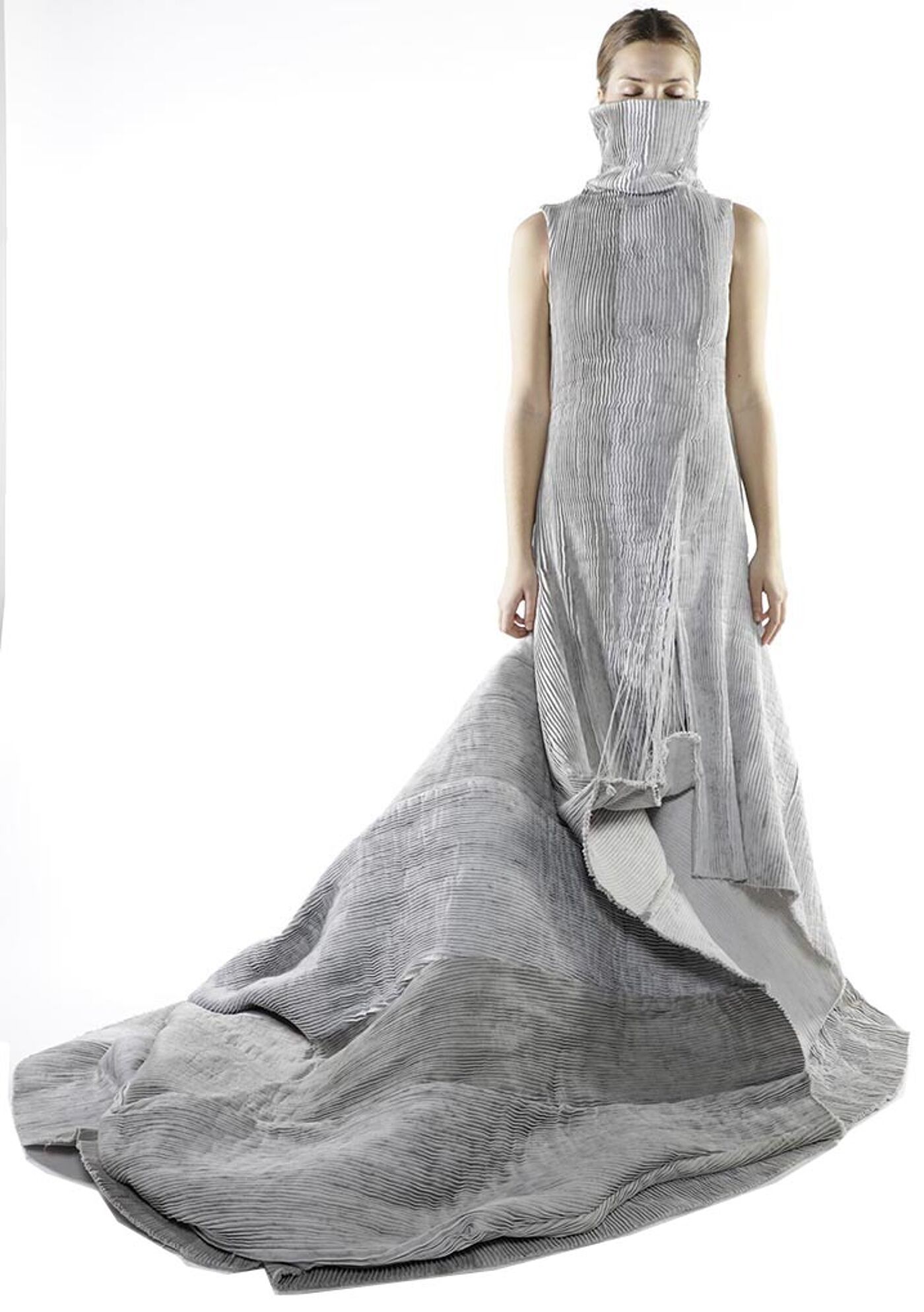 A woman in a long, pleated, grey dress with a high turtleneck stands against a white background, the dress pooling dramatically around her feet.