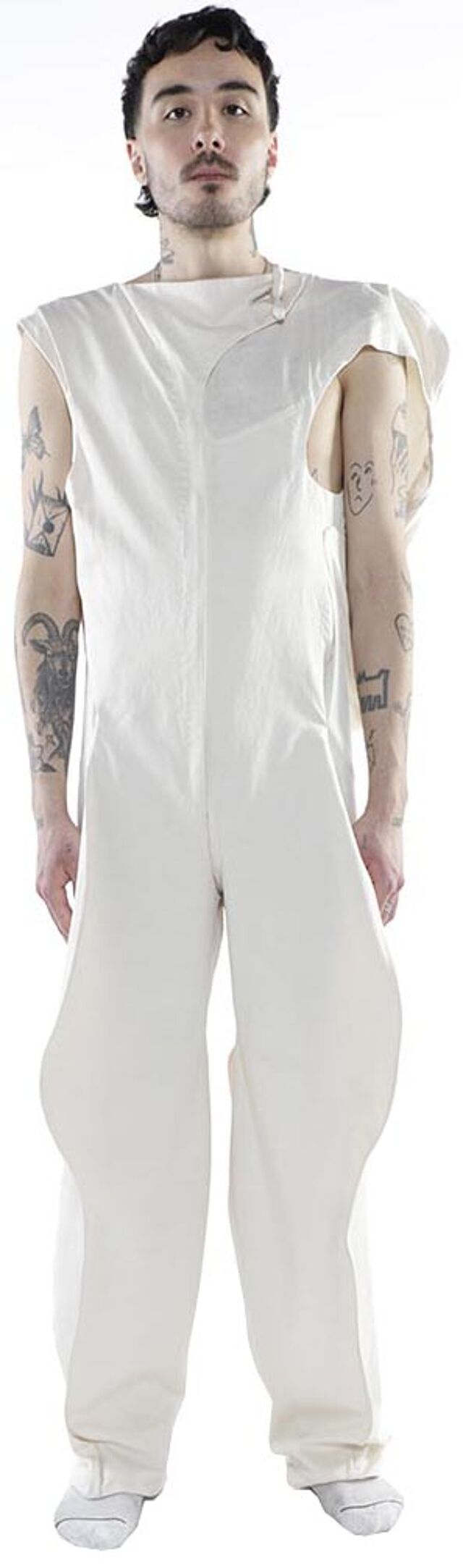 A person with tattoos on their arms is modeling a white sleeveless jumpsuit with a unique cut and is wearing white socks.