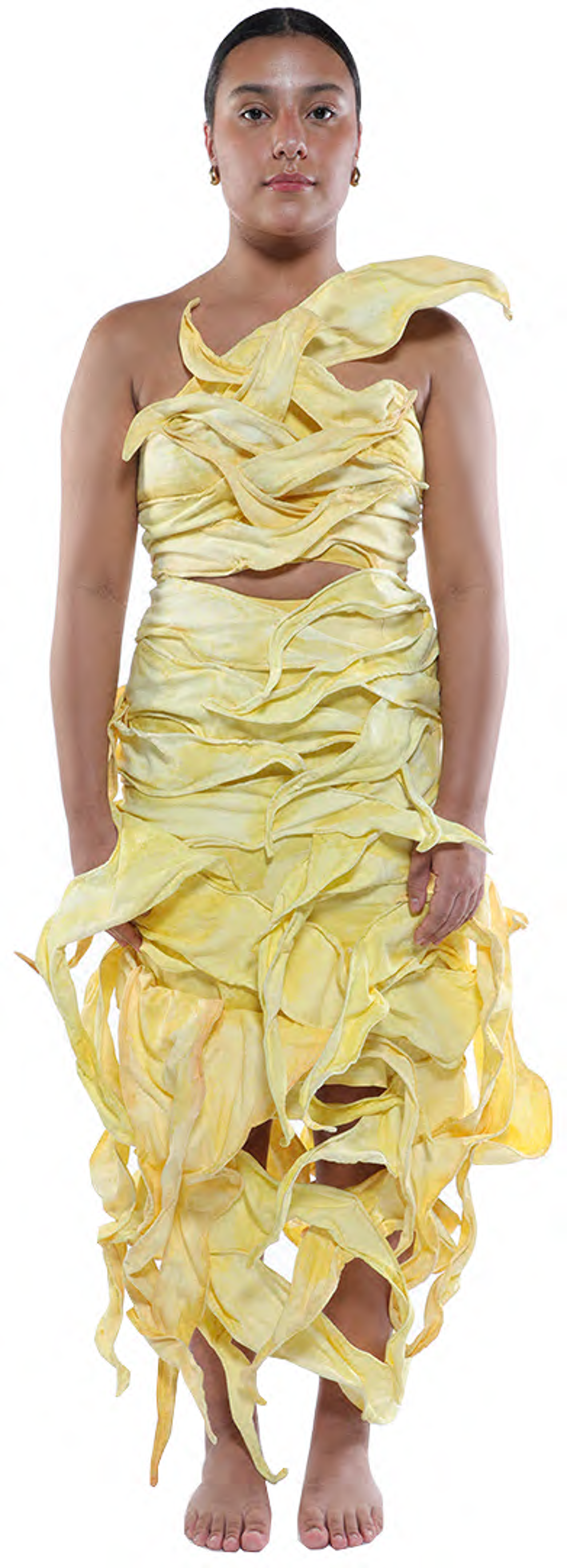 A young woman with tan skin and dark hair stands against a white background wearing a flowing, asymmetrical, pale yellow dress that appears to be made of fabric draped in layers.