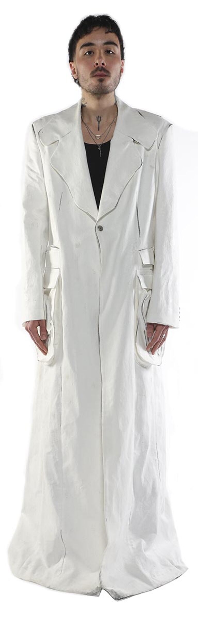A young man with short dark hair and tattoos stands against a white background, wearing a long, white coat with pockets and a collared shirt underneath.