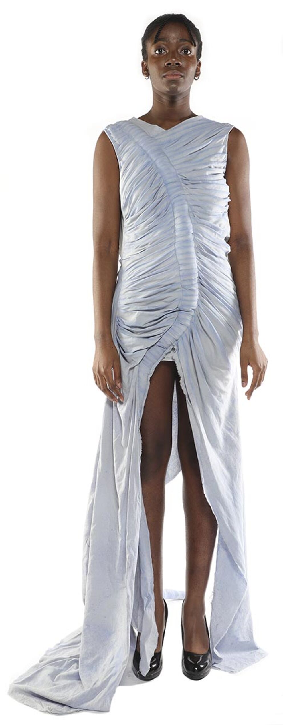 A slender Black woman with dark hair pulled back stands facing the camera in a light blue, draped maxi dress with a high slit, wearing black heels.