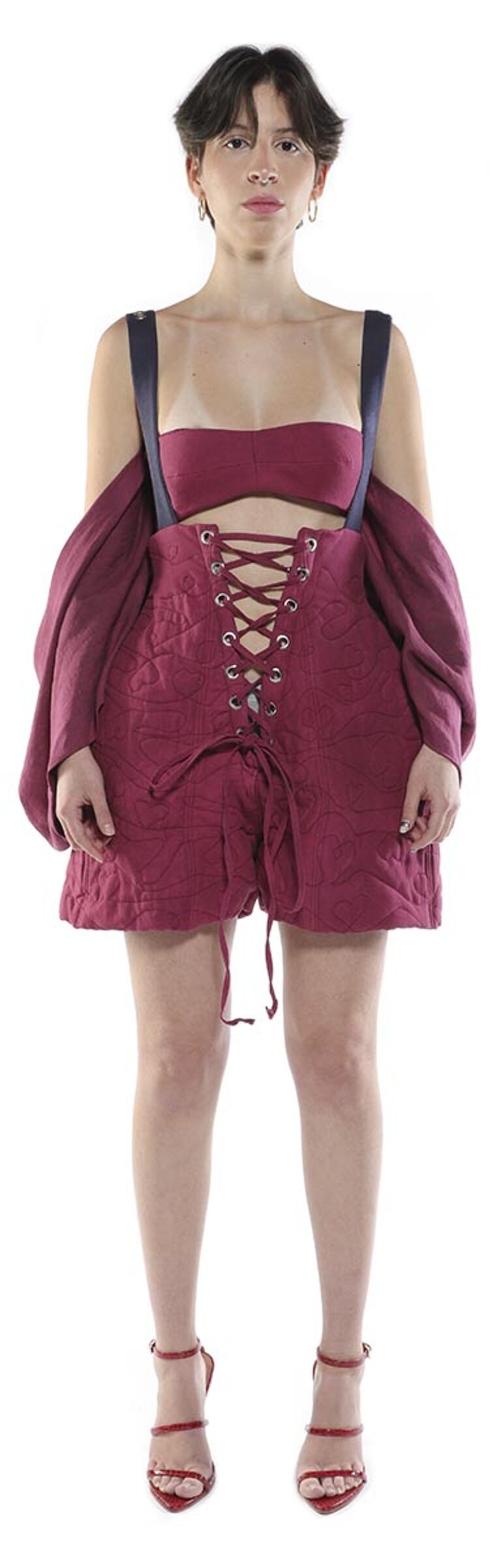 A woman with short dark hair is modeling a maroon corset-style top and shorts with dark straps, paired with red high-heeled sandals.