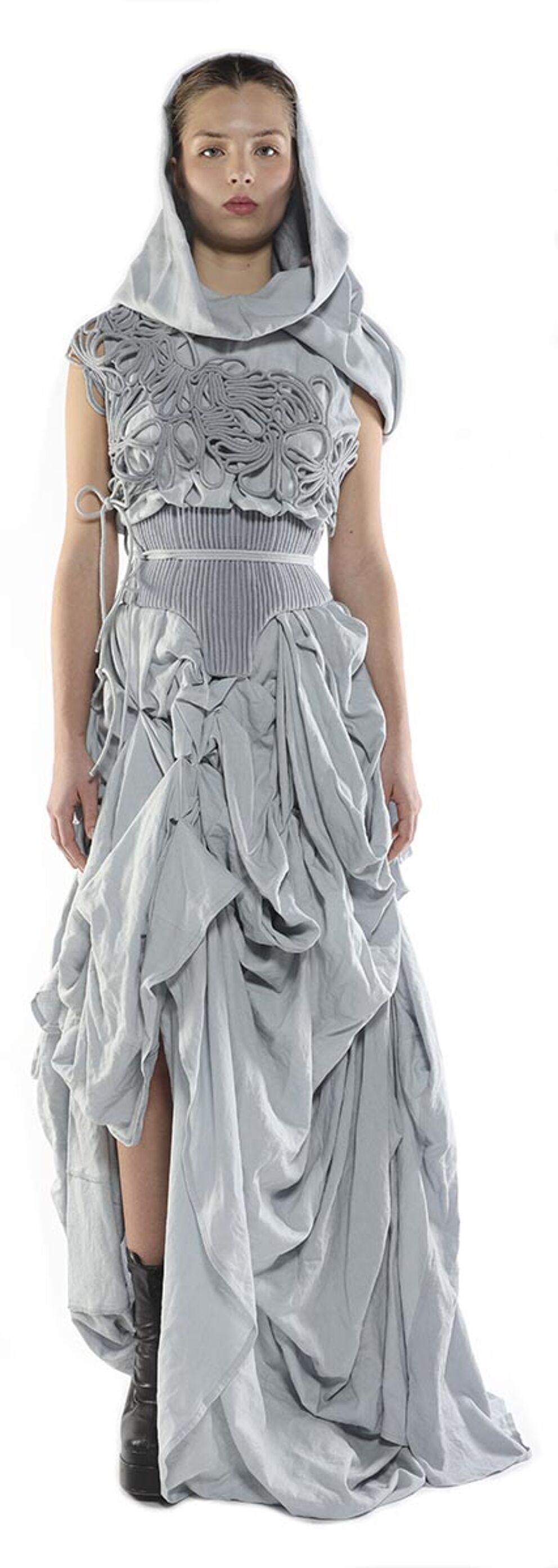A model wears a flowing, light-grey, asymmetrical gown with a hooded neckline and intricate bodice detail, paired with dark boots.