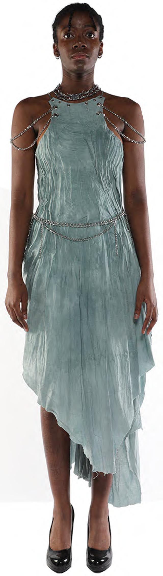 A dark-skinned person with short hair stands wearing a light teal asymmetrical dress adorned with silver chains and black high heels.