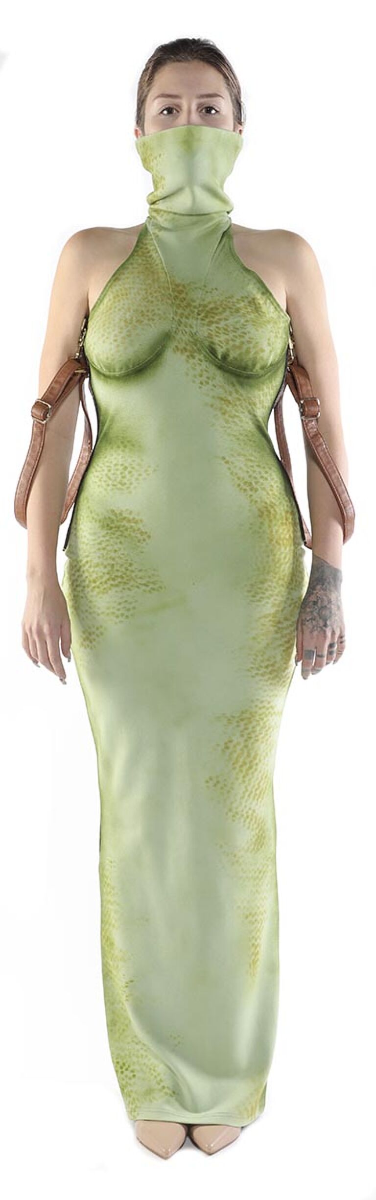 A woman in a pale green, form-fitting dress with a high neck and straps, and a matching face covering, stands against a white background.