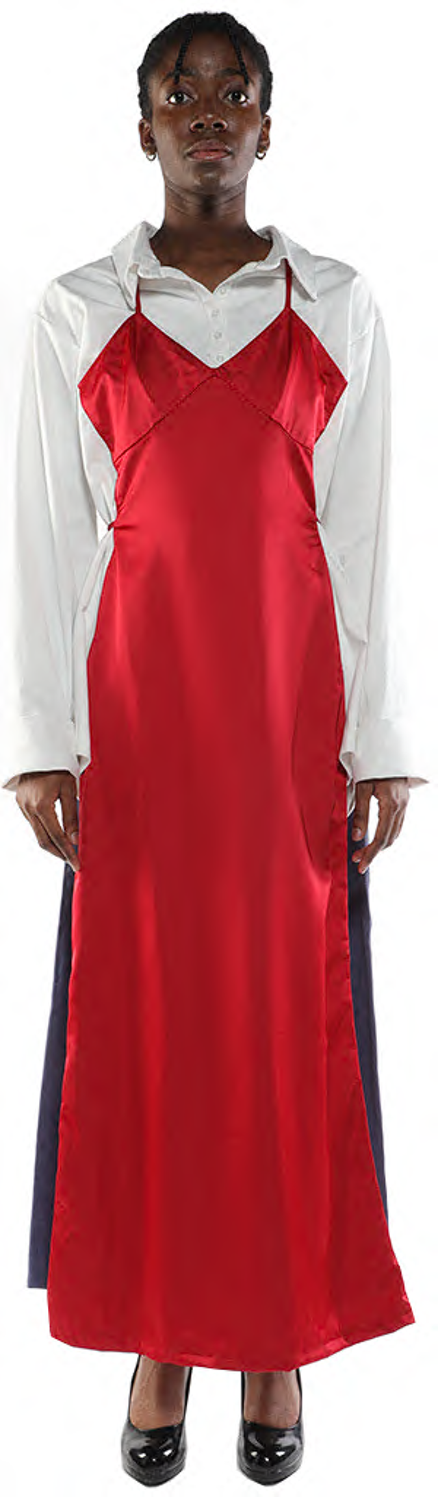 A person with dark skin and short black hair wears a long red satin slip dress over a white collared shirt and dark pants, and black heels.