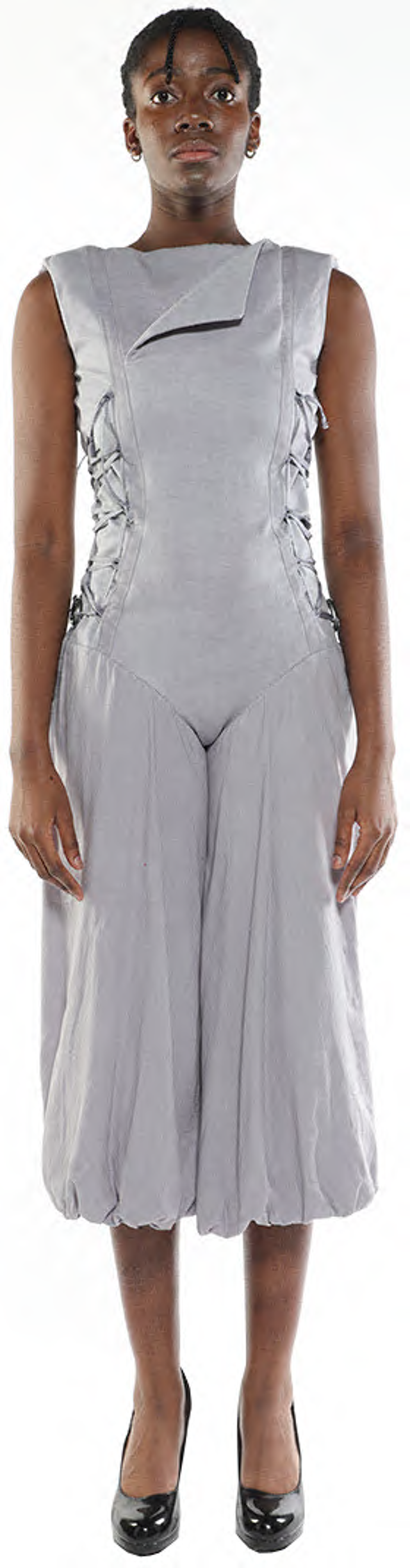 A dark-skinned model with short black hair wears a light gray sleeveless jumpsuit with a unique laced bodice and balloon-style pants, paired with black high heels, posing against a white background.