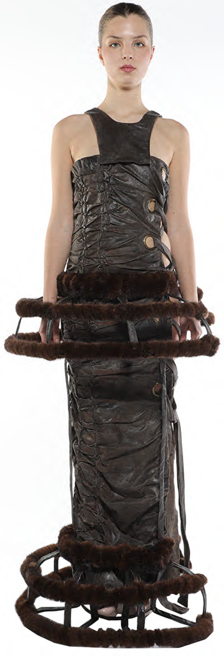 A slender woman with her hair pulled back stands facing forward, wearing a dark brown, sleeveless, textured top and matching long skirt, both adorned with fur trim and metallic accents.