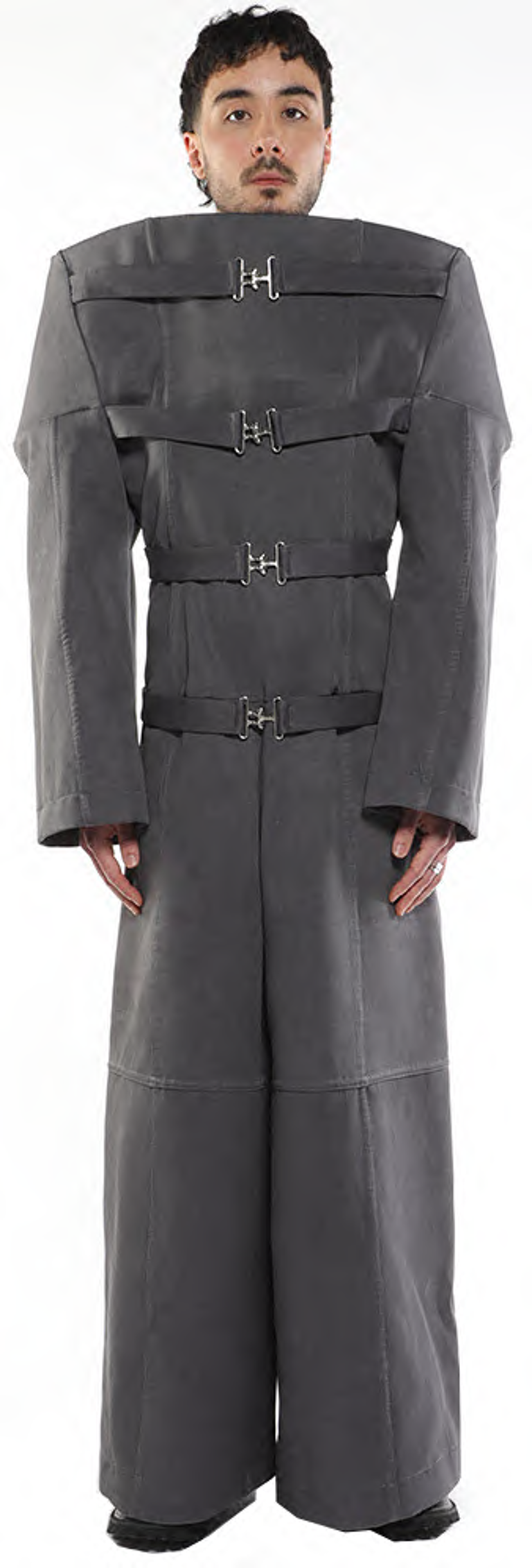 A person with short brown hair is standing and wearing a long, gray, loose-fitting coat with several straps and buckles.
