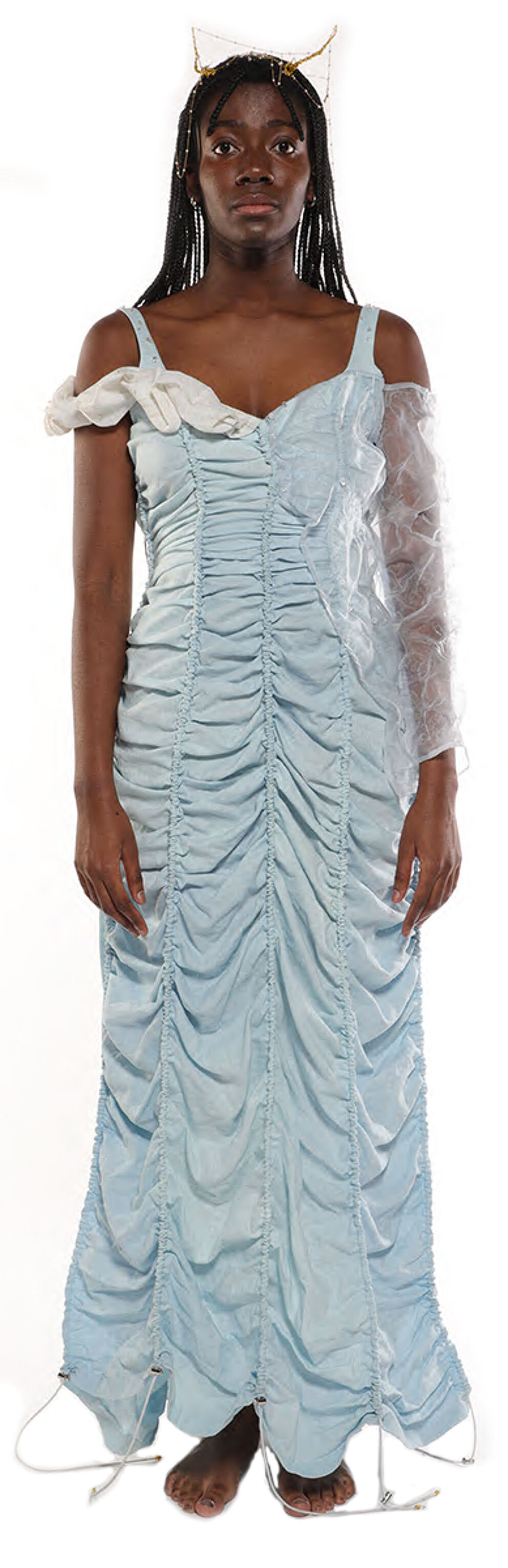 A light blue, ruched maxi dress with a sheer overlay on one sleeve is worn by a dark-skinned woman with long black braids and a crown.