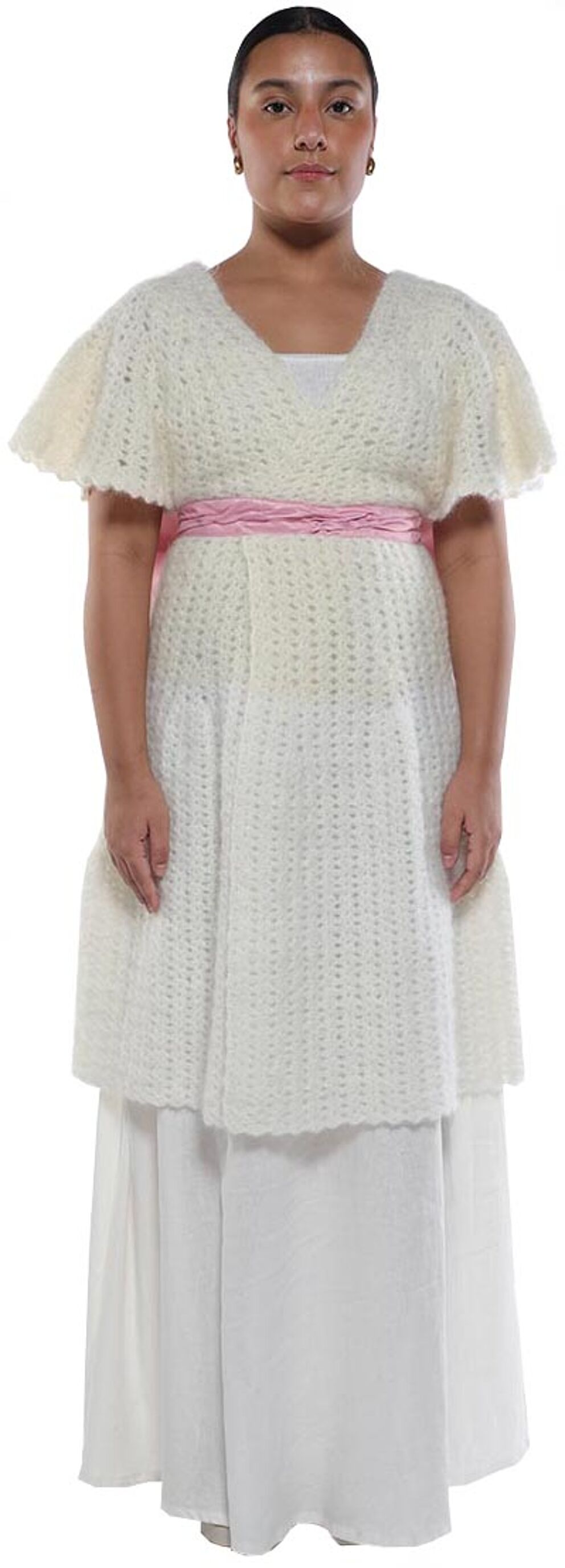 A woman with light skin and dark hair is wearing a long, white, crocheted dress with a pink sash.