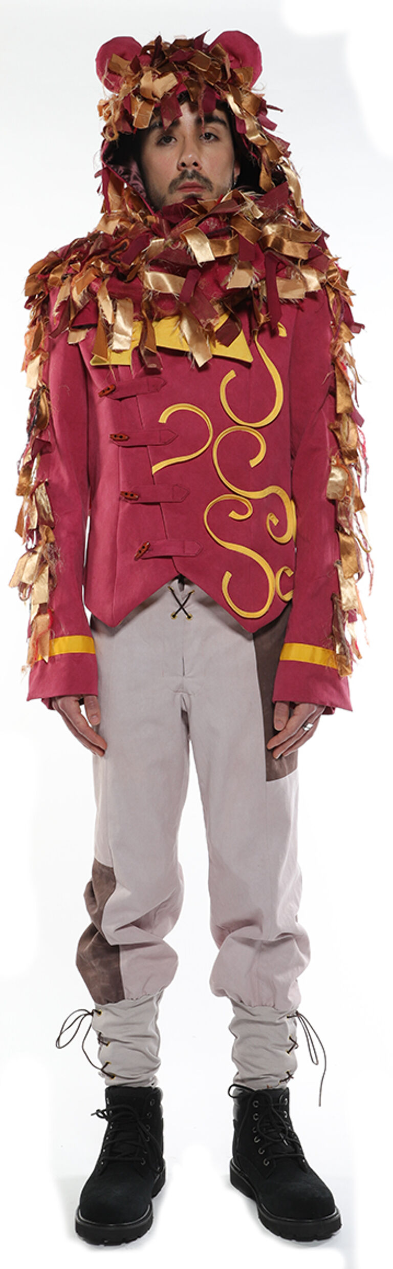 A person wearing a red jacket with gold embroidery, light-grey pants, brown boots, and a lion-mane-like hood stands against a white background.