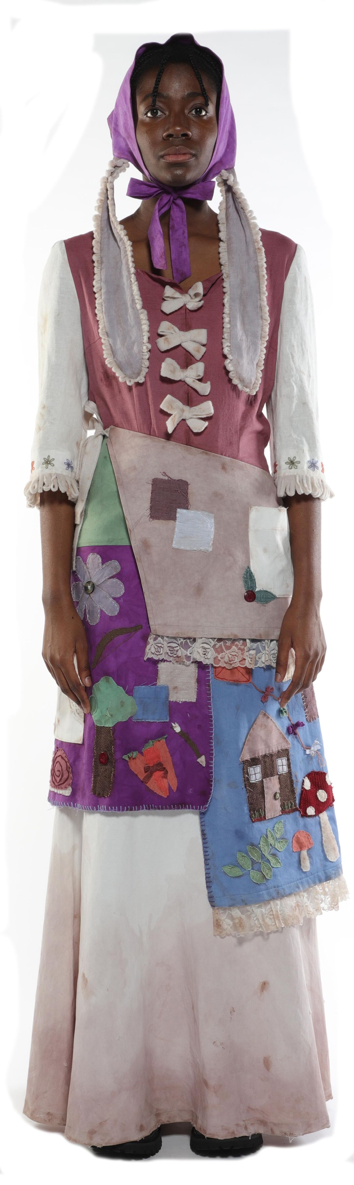 A young Black woman in a patched, multi-colored dress and purple bonnet stands against a white background.
