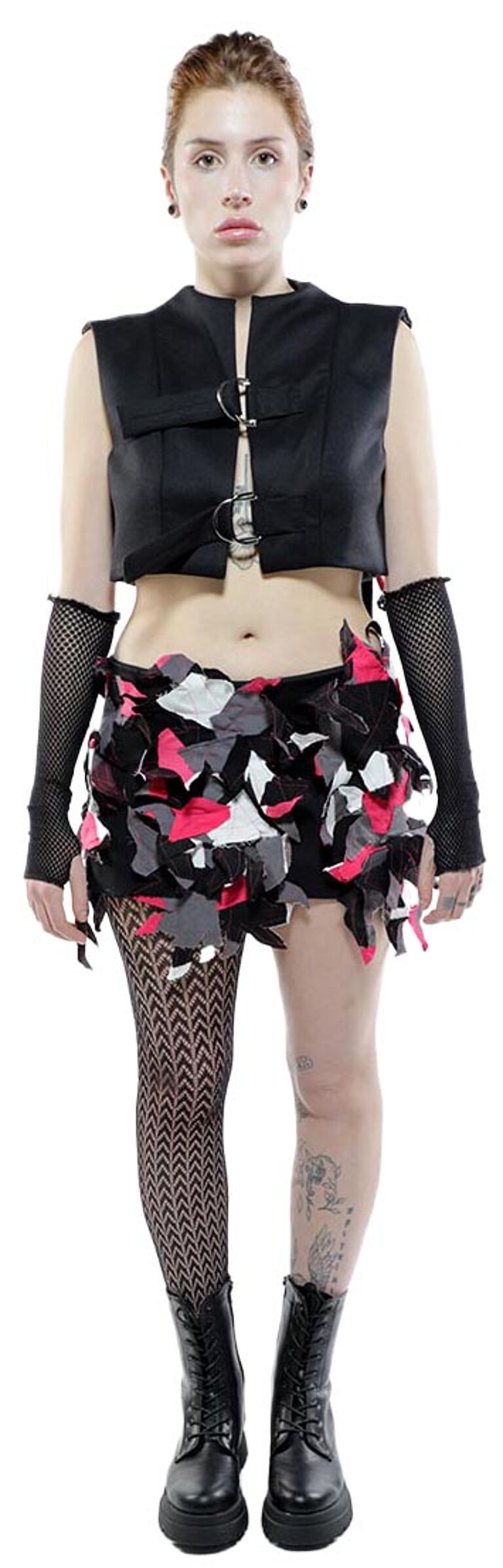 A young woman with red hair is modeling a black cropped vest, a multicolored short skirt, fishnet stockings, and black combat boots.