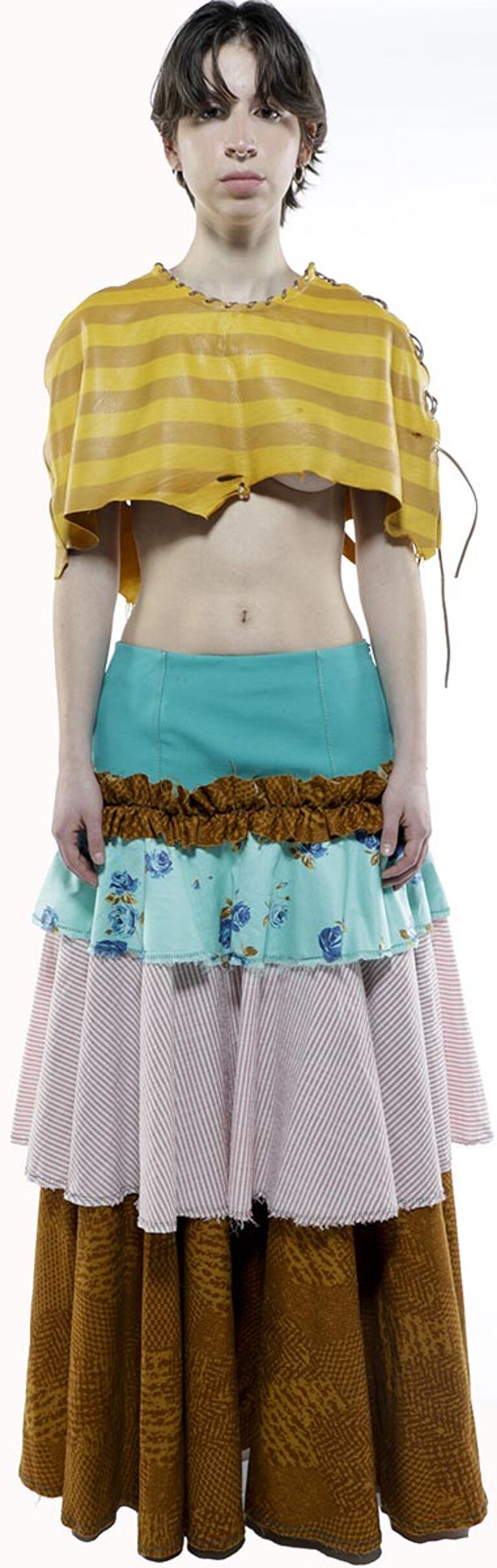 A person with short brown hair wears a mustard-yellow striped cropped top and a layered skirt with teal, floral, pink, and brown sections.