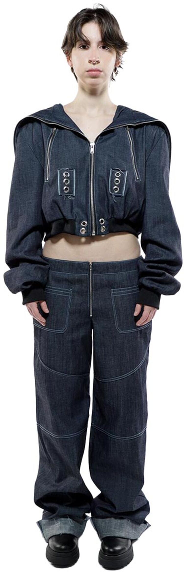 A young woman with dark hair models a dark denim hooded jacket and matching pants, both featuring decorative stitching and metal accents.