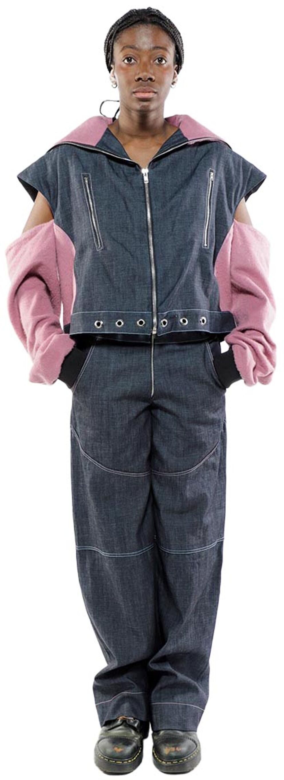 A person with dark skin and a dark head wrap wears a denim jumpsuit with pink sleeves and dark shoes.