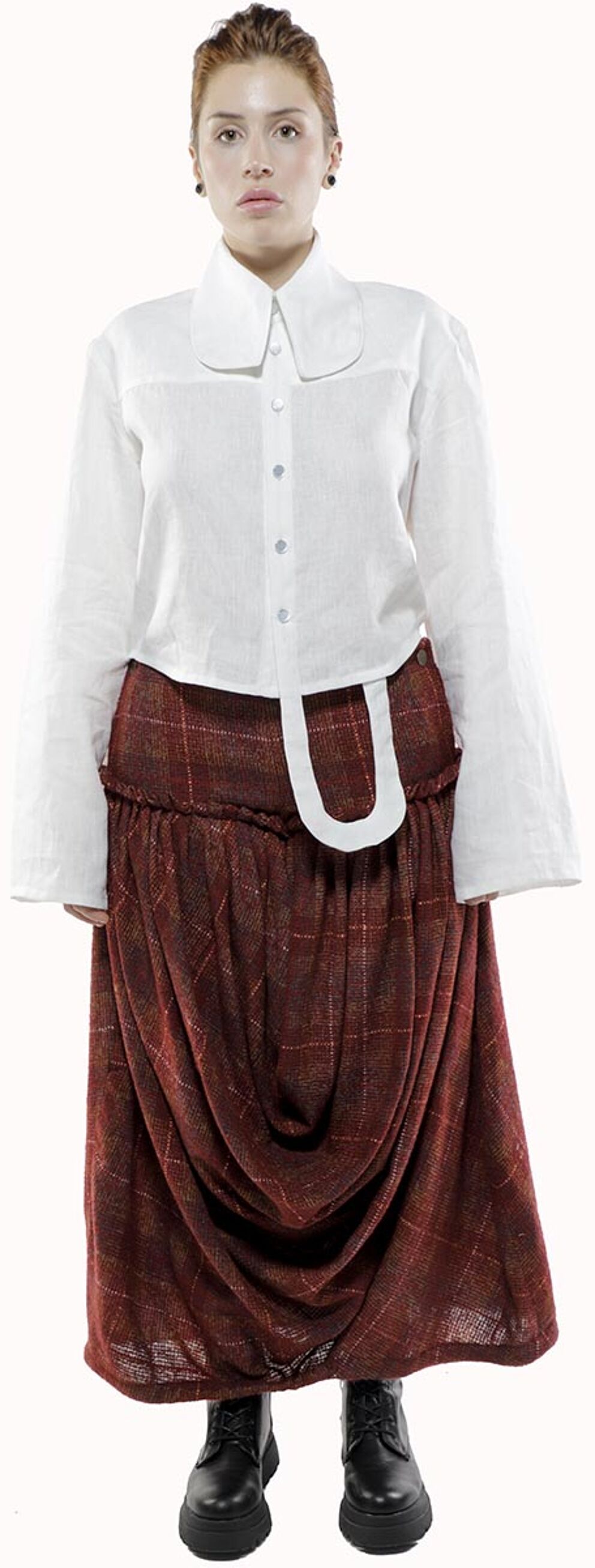 A woman with reddish-blonde hair wears a long-sleeved white linen shirt over a gathered maroon plaid skirt and black boots.
