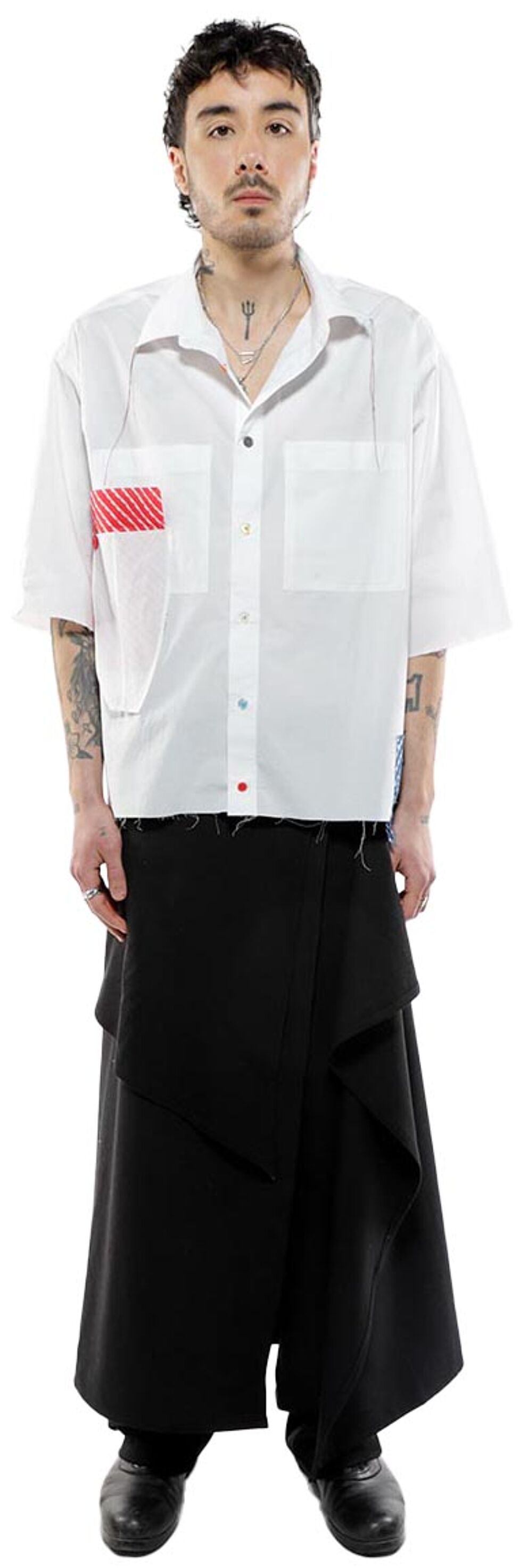 A young man with dark hair and tattoos stands against a white background, wearing a white short-sleeved button-down shirt with a red and white striped patch on the left breast pocket, and a black asymmetrical wrap skirt.