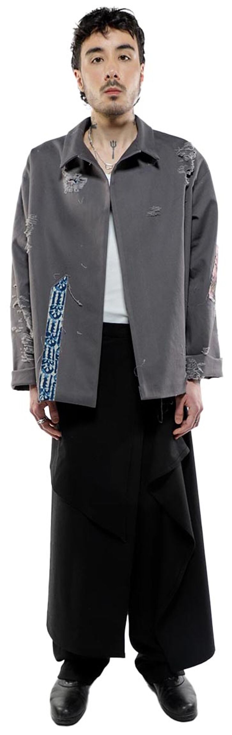 A young man with dark hair and a neutral expression stands against a white background, wearing a gray, distressed jacket over a black, draped skirt and dark shoes, accessorized with a thin necklace.