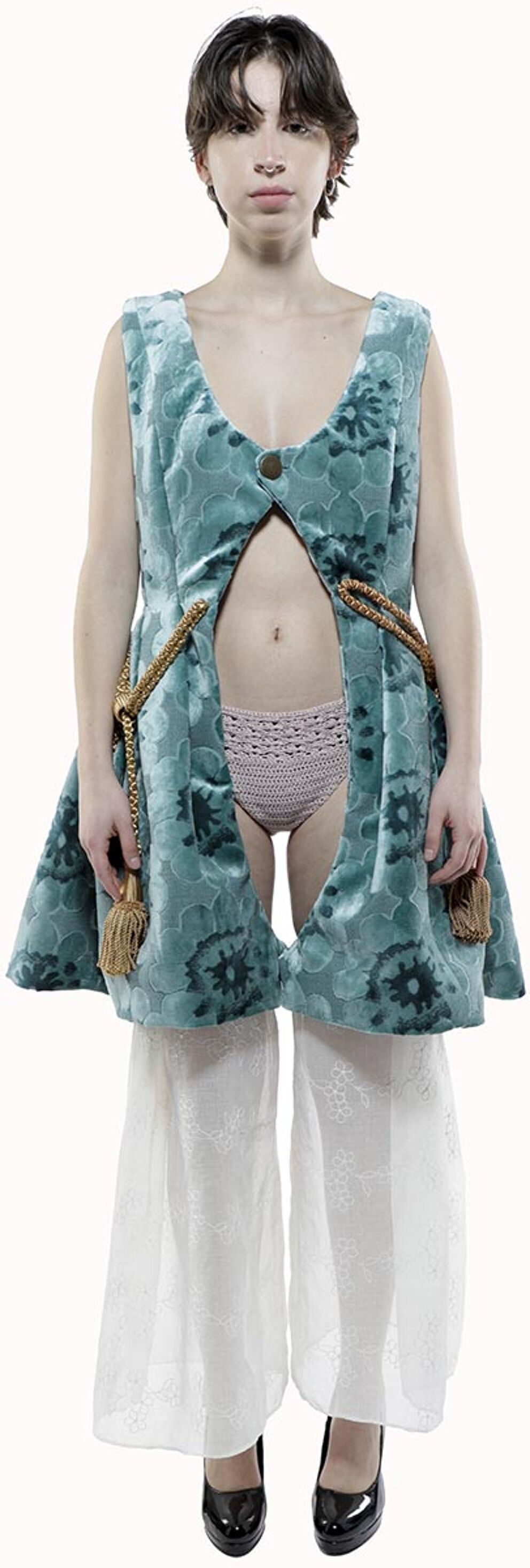 A person with short brown hair wears a teal velvet vest over a pale pink crocheted underwear and white pants, accessorized with gold tassels.