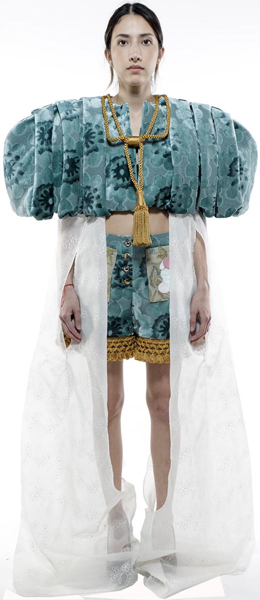 A woman with dark hair, wearing a teal patterned top with puffy shoulders, matching shorts, and a long white sheer overlay, stands against a white background.