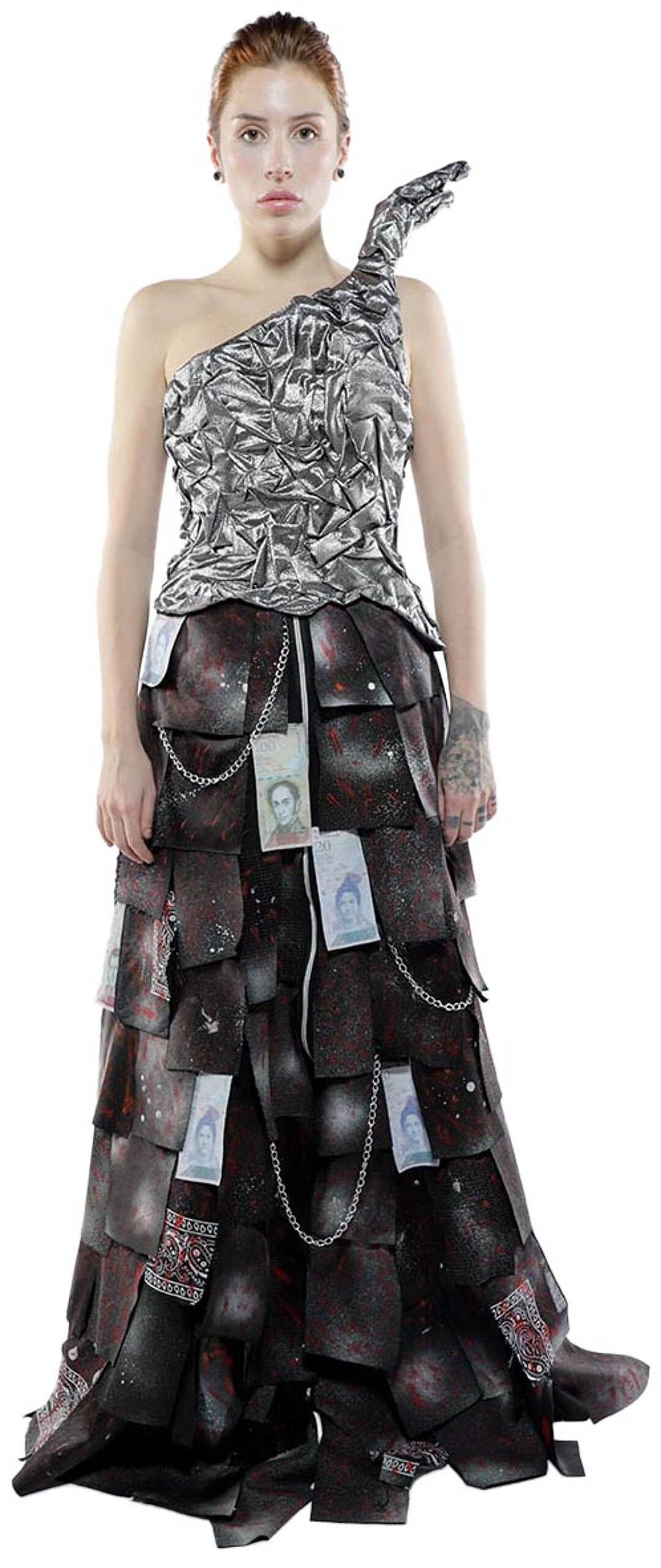 A woman with red hair models a long, asymmetrical dress with a silver bodice and a dark, layered skirt decorated with chains and what appears to be currency.