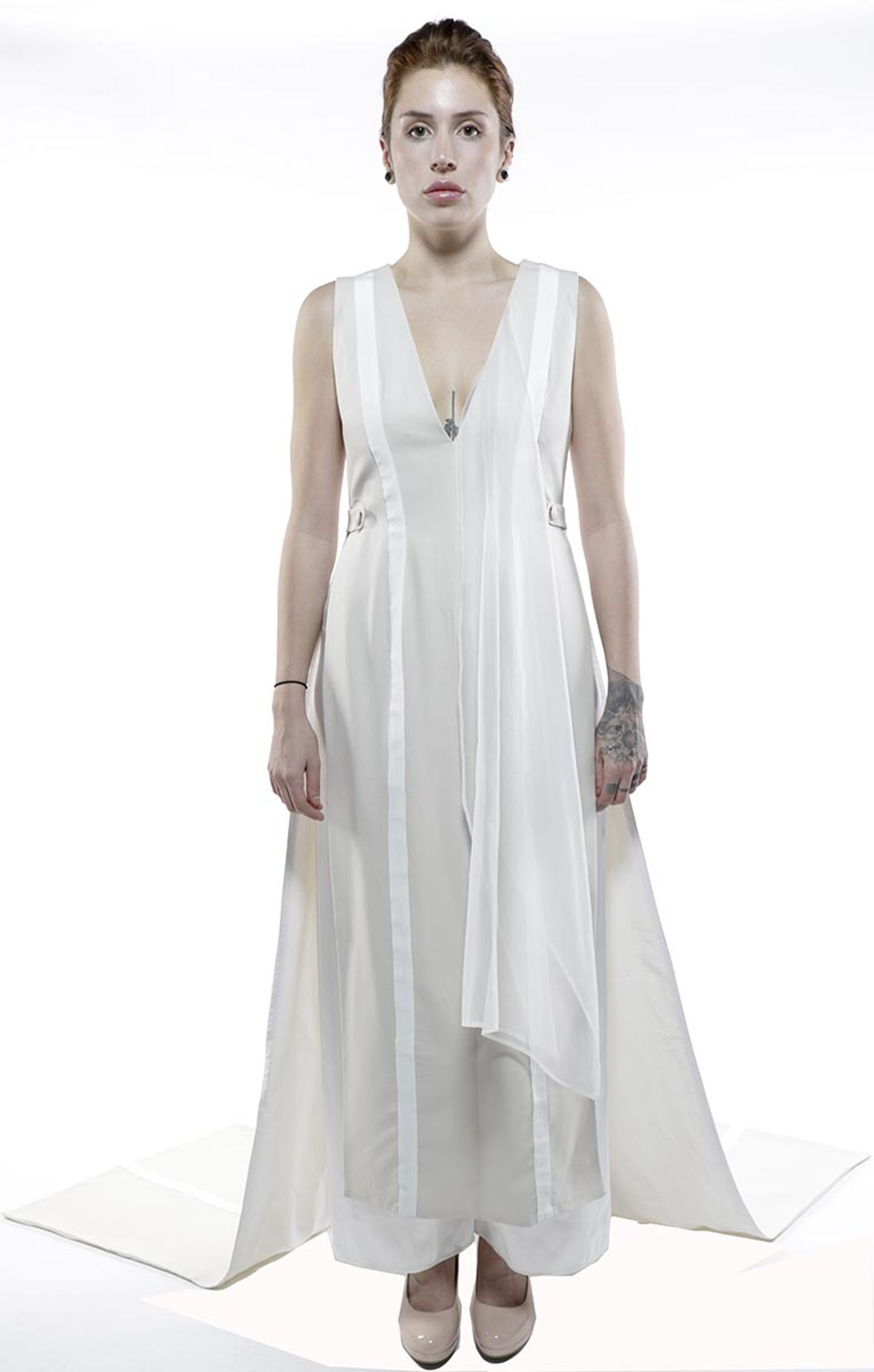 A woman with red hair and tattoos stands against a white background wearing a long, white, sleeveless dress with a flowing train.