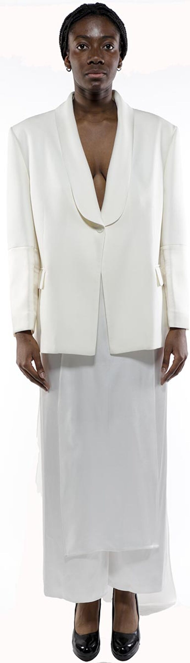 A dark-skinned woman with dark hair styled in a bun wears an off-white blazer and matching long skirt, accessorized with black shoes and earrings, standing against a white background.