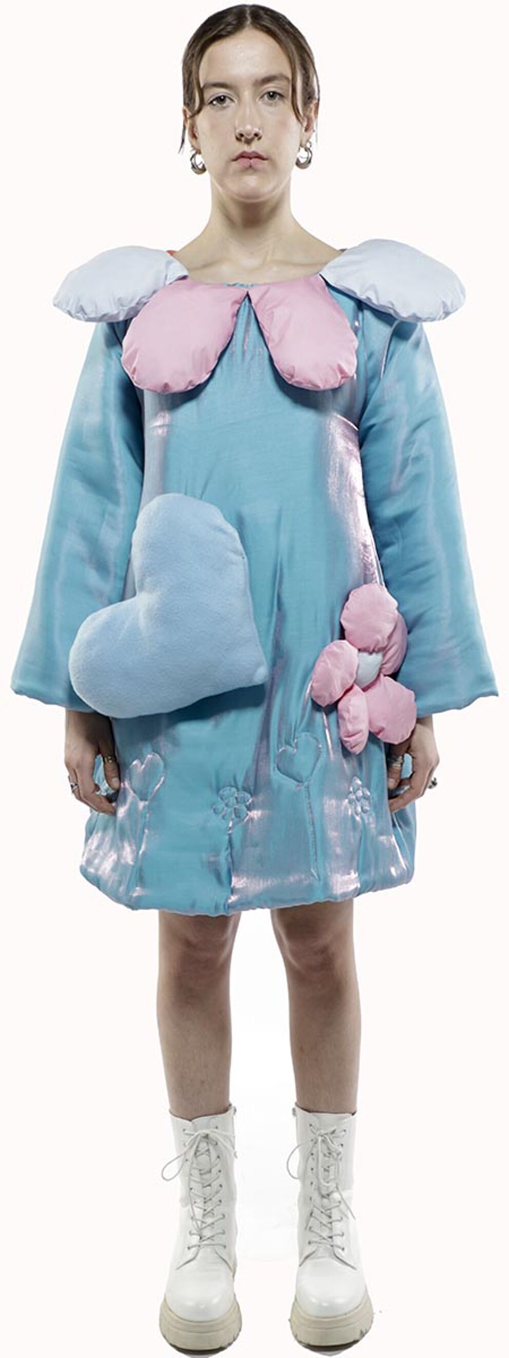 A young woman with shoulder-length brown hair wears a light blue dress adorned with pink and light blue fabric hearts and flowers, complemented by white lace-up boots.