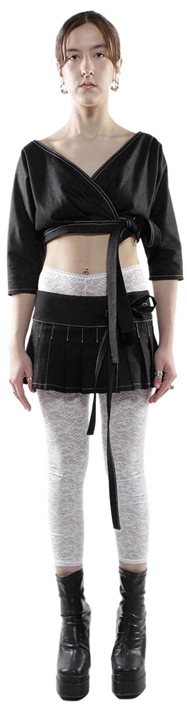 A young woman with shoulder-length brown hair is modeling a black wrap top, a black pleated mini-skirt, light gray leggings, and black platform boots.