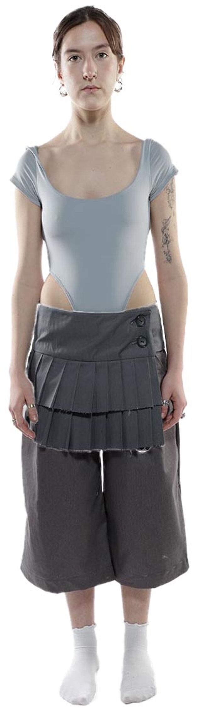 A young woman with shoulder-length brown hair is modeling a light-grey bodysuit and a layered grey pleated skirt and matching shorts.