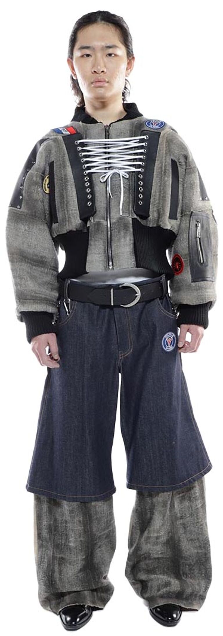 A person with dark hair styled back, wearing a gray, cropped jacket with black accents, a black belt, and dark denim pants with long, gray fabric overlays, stands against a white background.