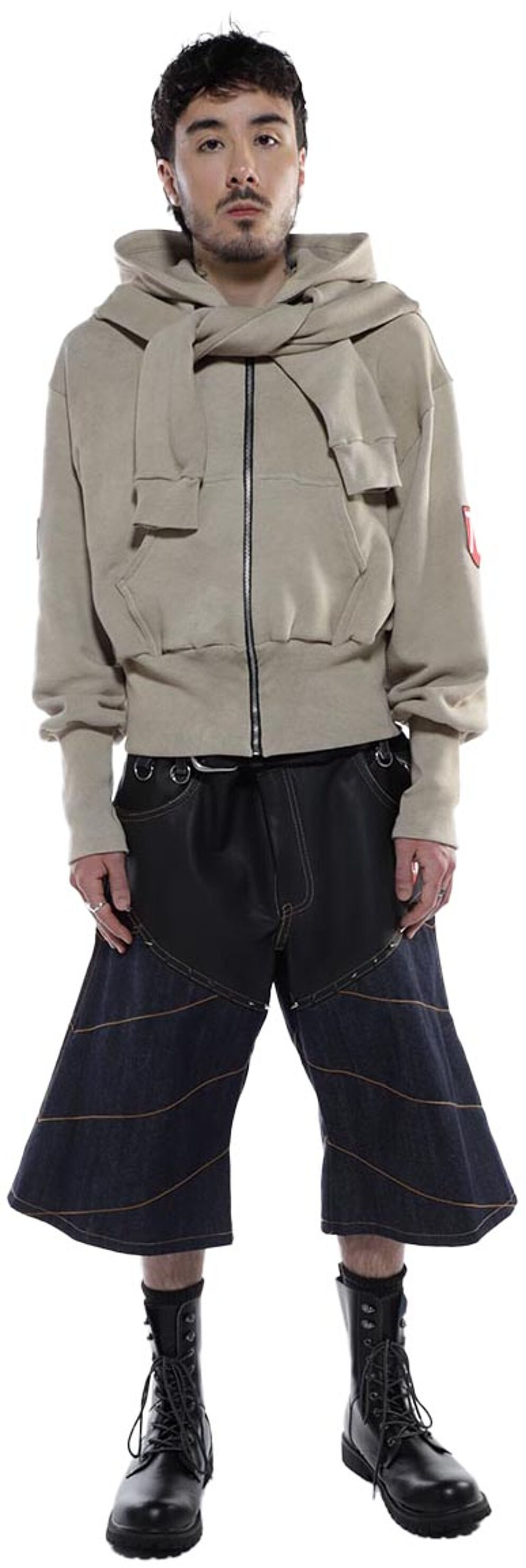 A person with short brown hair wears a beige zip-up hoodie with a scarf-like collar, dark-colored shorts with decorative stitching, and black combat boots.