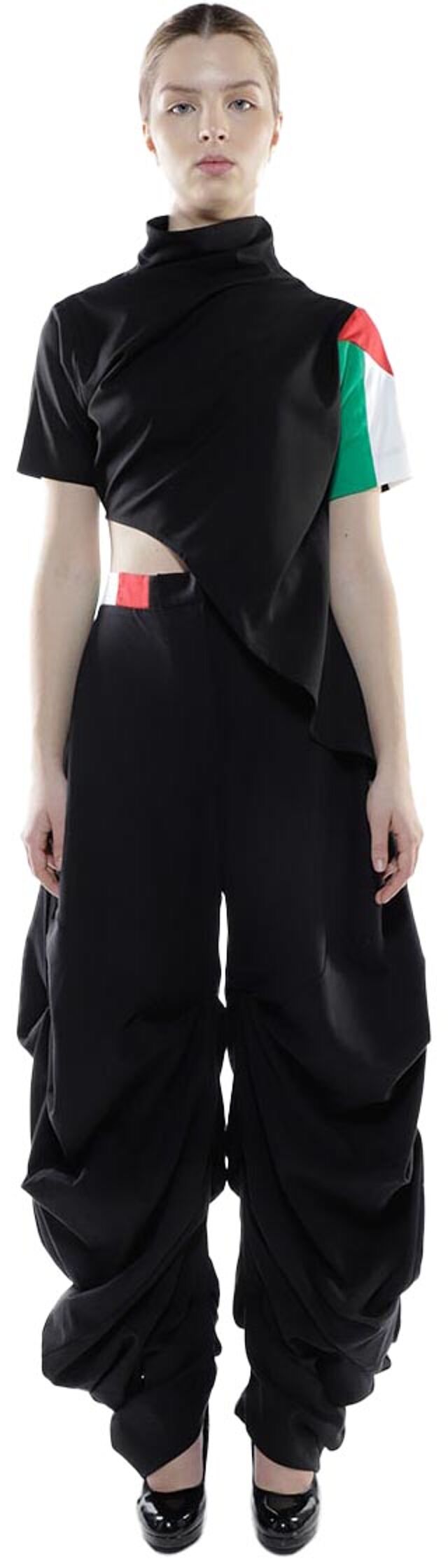 A woman with light skin and dark hair is modeling a black asymmetrical top with a color-blocked sleeve and matching wide-legged pants.