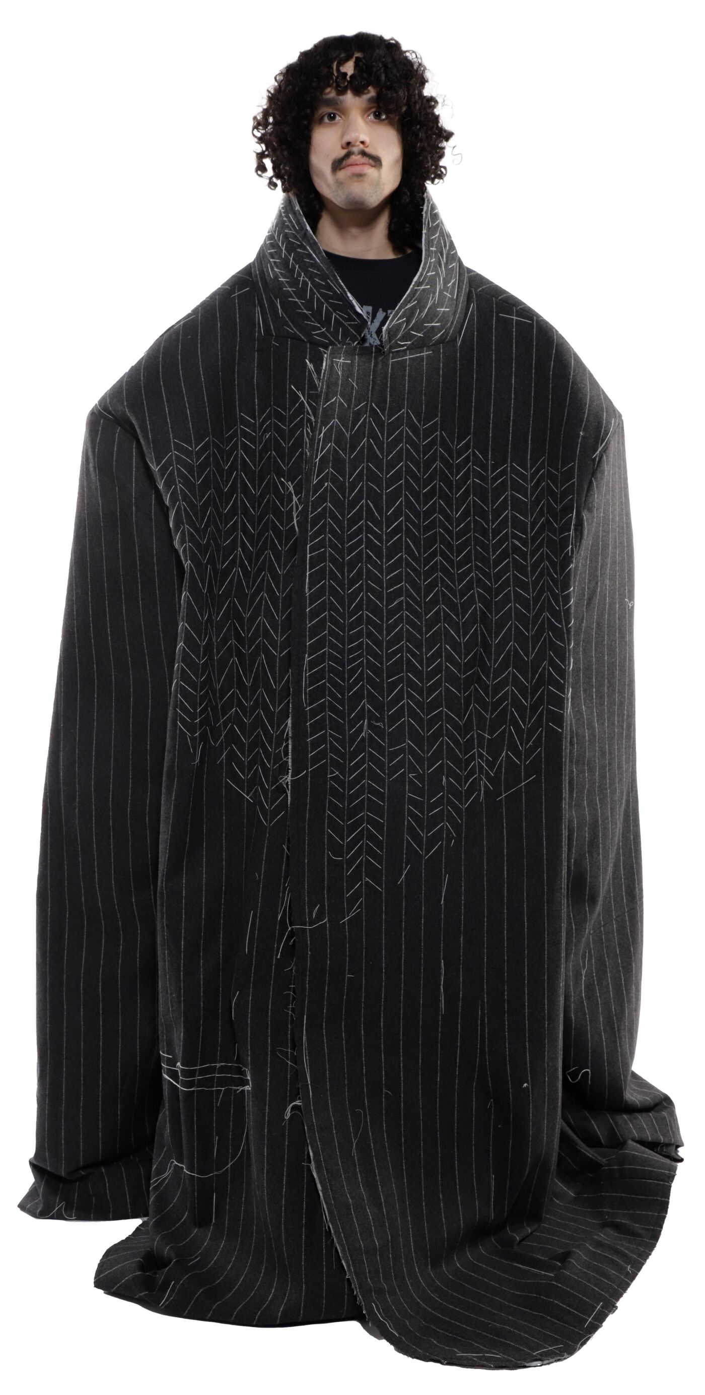 A person with curly hair wears an oversized, dark-striped, long coat with a high collar.