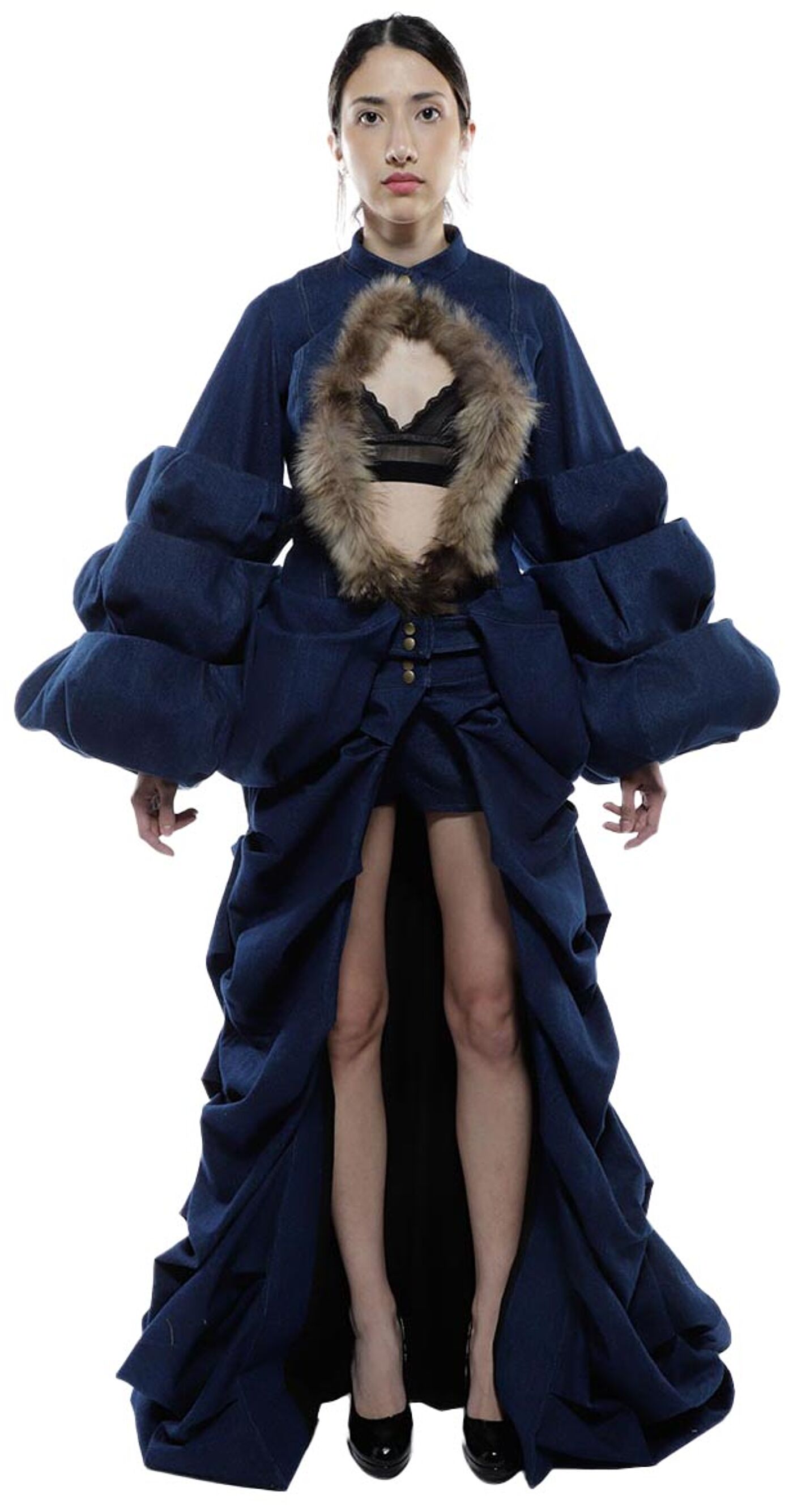 A woman in a dark blue denim dress with large puffy sleeves and a fur collar stands with her legs slightly apart and her hands at her sides.