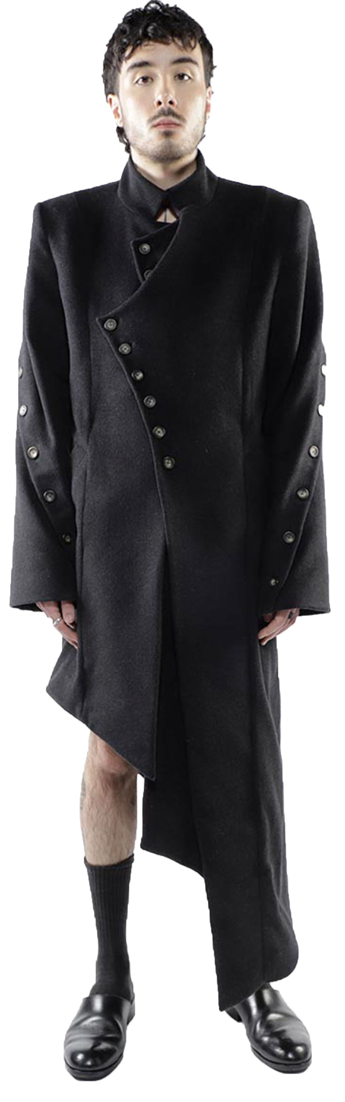 A person with short dark hair is wearing a long, black, asymmetrical coat with buttons and dark socks and shoes.