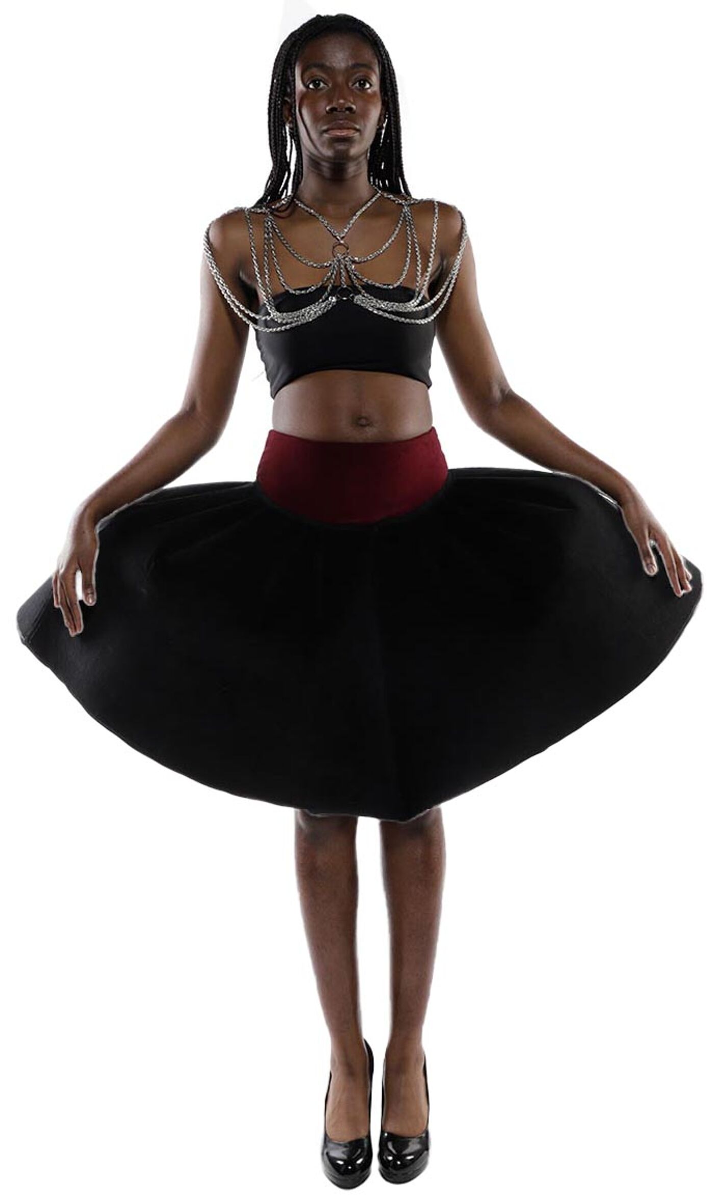 A dark-skinned woman with long black hair and a silver body chain wears a black skirt with a maroon waistband and black high heels.