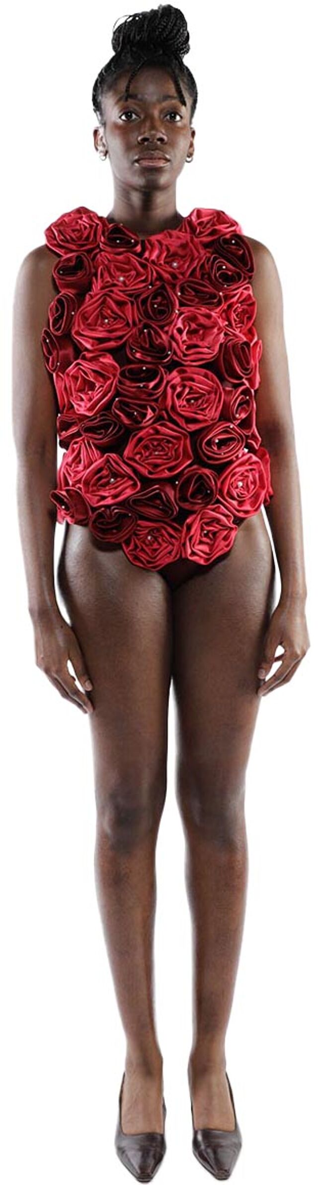 A dark-skinned woman with dark hair styled in a bun wears a form-fitting, sleeveless red bodysuit adorned with numerous fabric rose appliqués and dark brown high-heeled shoes.