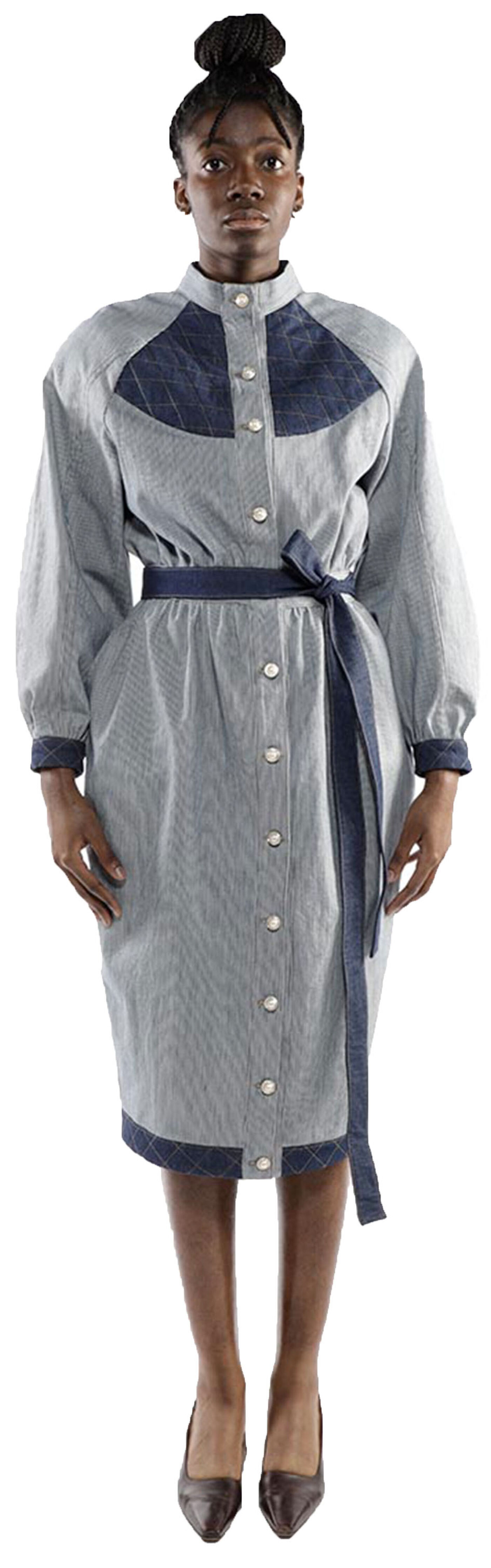 A dark-skinned woman with her hair in a bun wears a long-sleeved denim dress with a contrasting yoke and a self-belt, standing against a white background with her legs slightly apart and wearing dark-brown high heels.