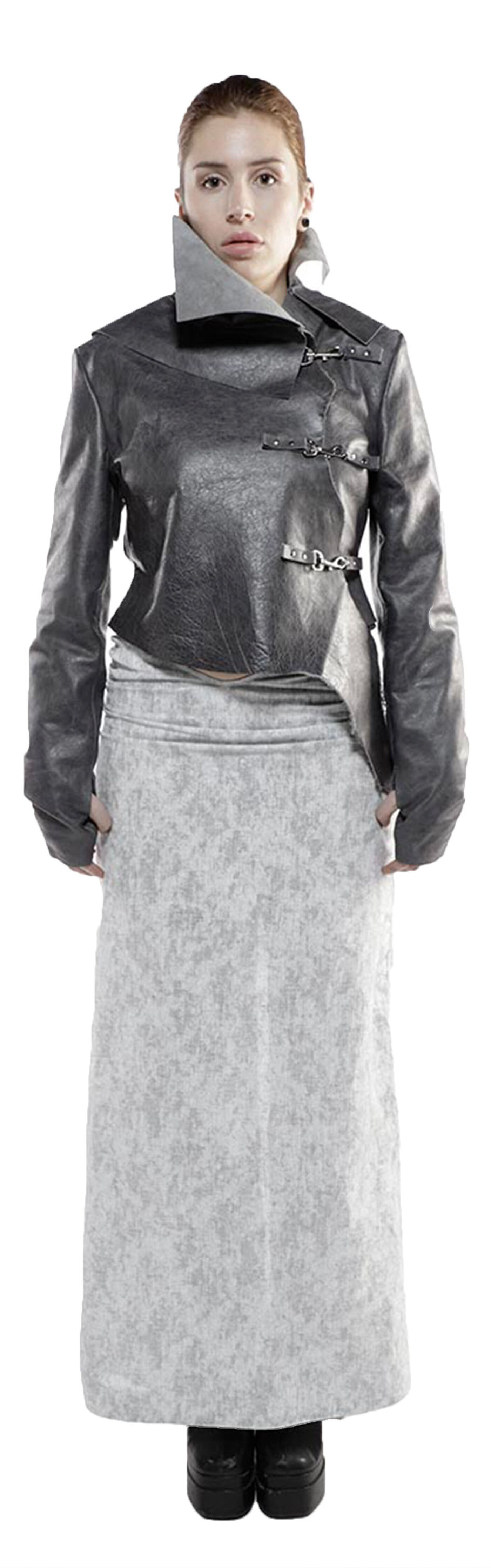 A woman with light skin and reddish-brown hair wears a dark gray leather jacket with a light gray high-collared shirt underneath, paired with a light gray, long, textured skirt and black boots.