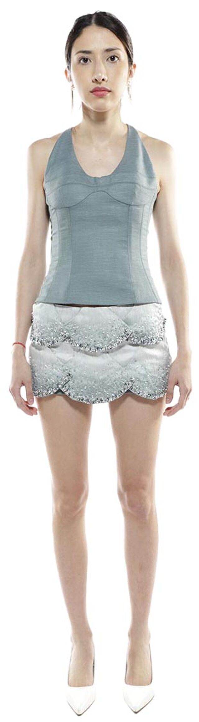 A slender woman with dark hair stands against a white background, wearing a light-blue halter top and a sequined, scalloped mini-skirt, and white high-heeled shoes.