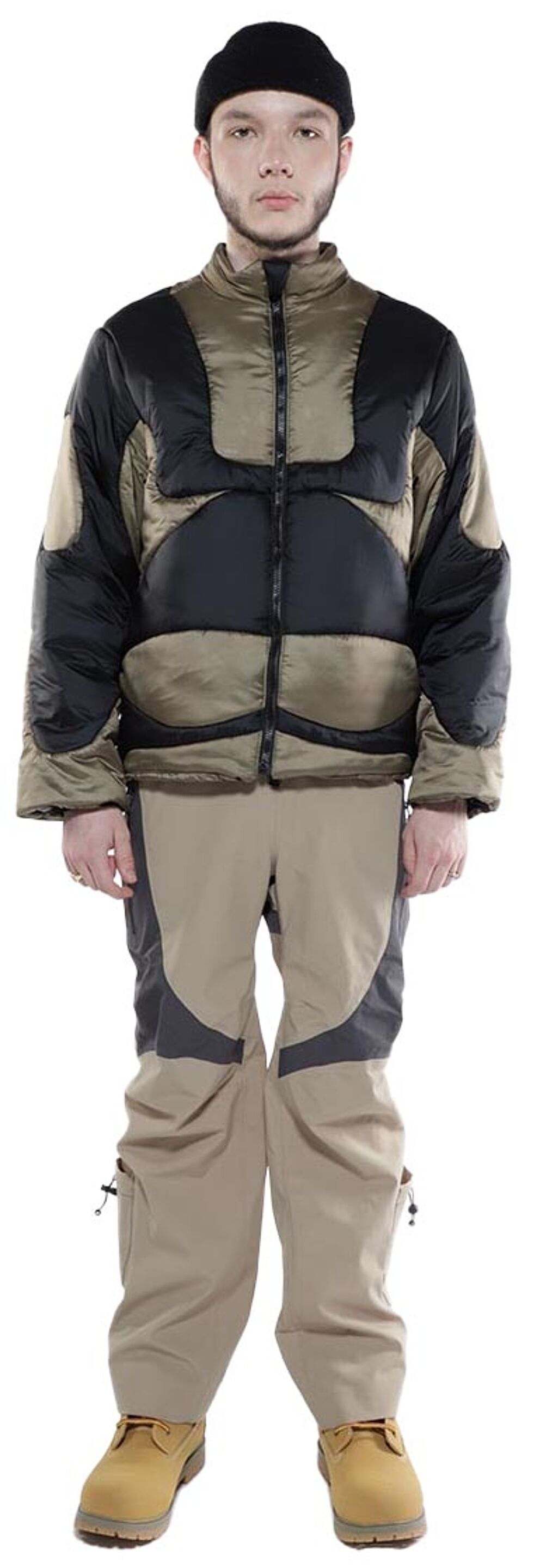 A man wearing a black and tan puffer jacket, tan pants with dark gray accents, and work boots stands against a white background.