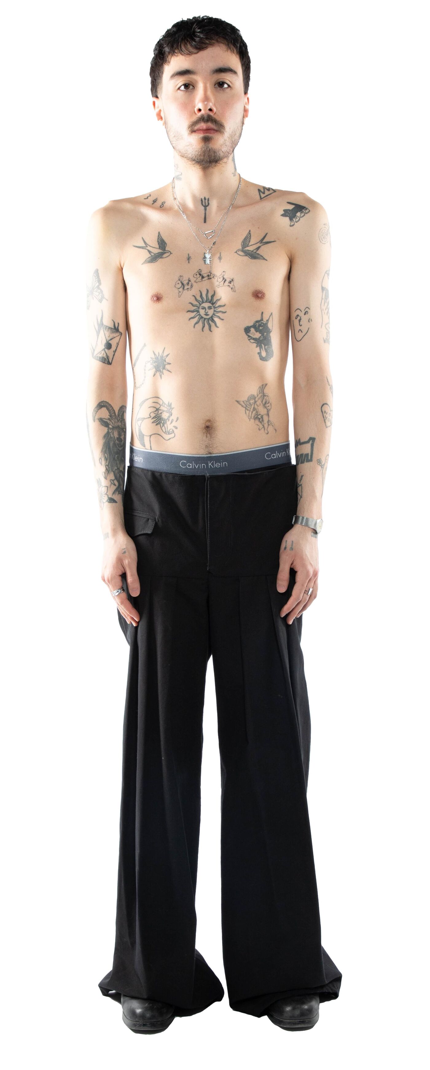 A slender, tattooed man with dark hair stands against a white background, wearing black wide-leg pants and black shoes, with a grey Calvin Klein underwear waistband visible.