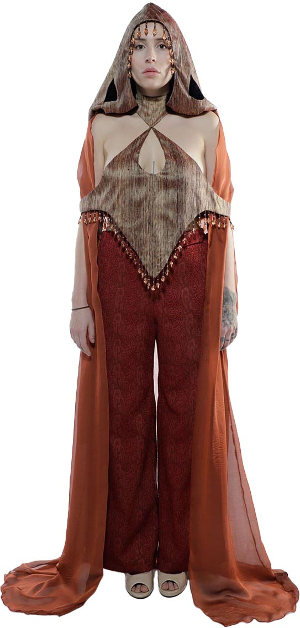 A woman in a flowing orange robe and patterned top with a hood adorned with beads poses against a white background.