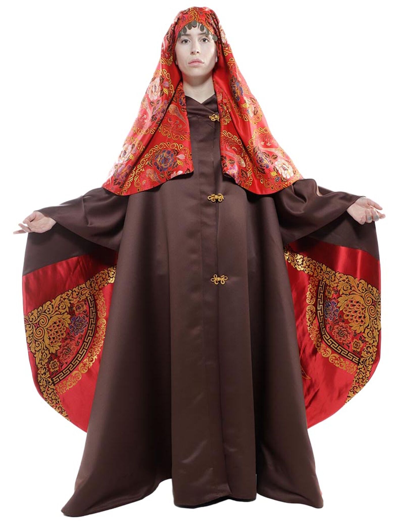 A woman in a long brown robe with red and gold patterned sleeves and head covering poses against a white background.